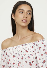 Haut blanc, dégageant les épaules, avec un motif floral en rouge et bleu. Comprend un col smocké et des manches amples et fluides. Texture de tissu léger.