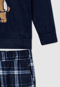Maglione in pile blu navy con design di un orso, abbinato a pantaloni da pigiama a quadri in blu navy, bianco e marrone, con elastico in vita.