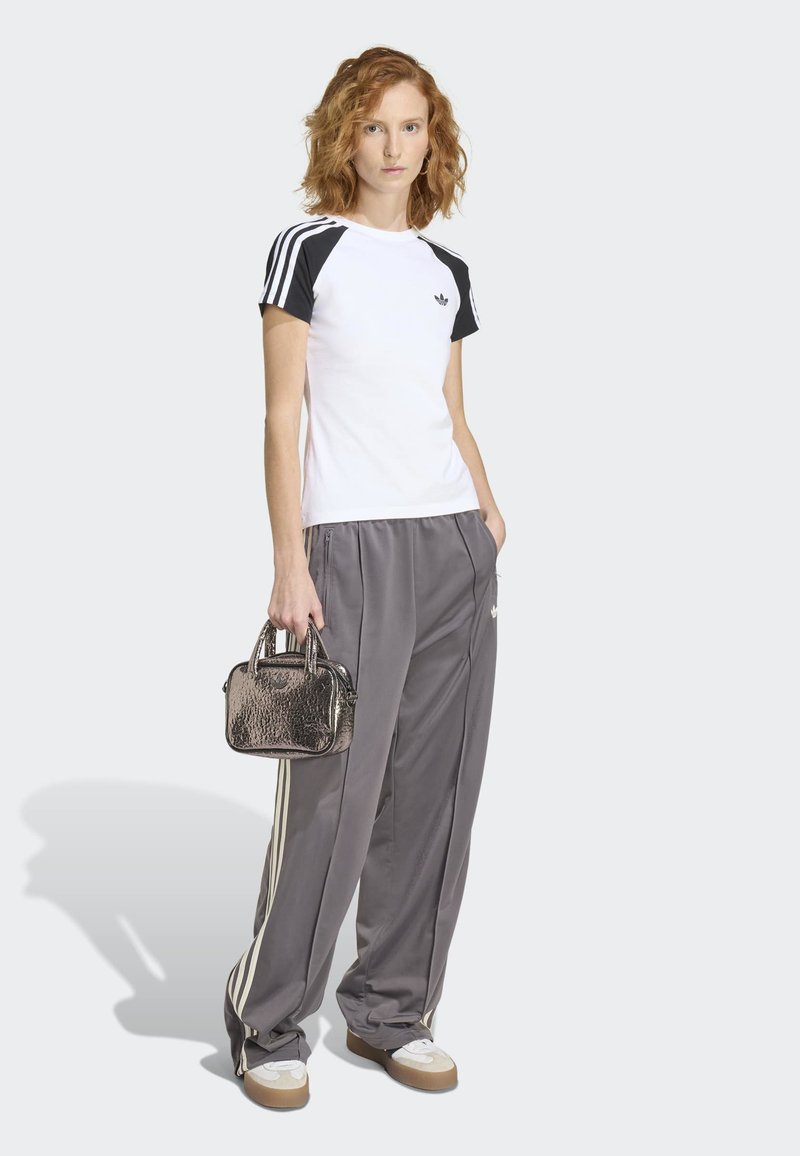 Femme aux cheveux roux bouclés portant un t-shirt Adidas blanc et noir, un pantalon de survêtement Adidas gris, des baskets blanches, tenant un petit sac à main métallique.