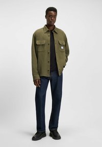 Olive green textured jacket with two chest pockets, black buttons, worn over a black shirt and paired with dark blue jeans and black shoes.