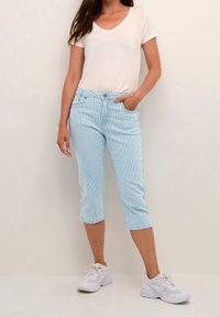 Světle modré pruhované capri kalhoty vyrobené z měkké tkaniny, v kombinaci s bílým tričkem s výstřihem do V a bílými sportovními botami.