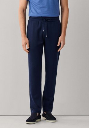 Hombre con pantalones azul marino con cordón, zapatos negros sin cordones y camiseta azul, de pie frente a un fondo gris claro.