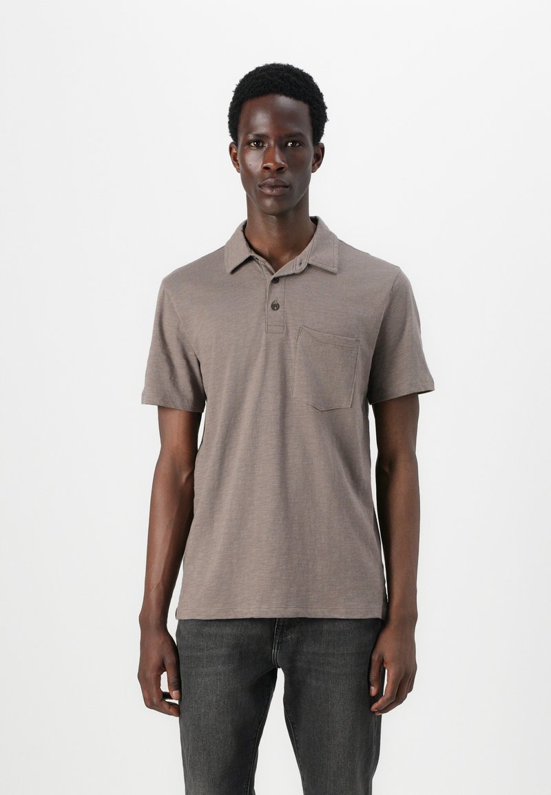 Polo shirt à manches courtes, marron, avec un tissu texturé, un col et une poche poitrine. Porté avec un jean gris foncé.