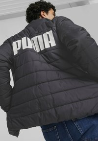 Giacca nera imbottita con dettagli cuciti e grande logo bianco "PUMA" sulla schiena. Il tessuto appare liscio con una leggera lucentezza.