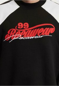Sweat-shirt noir avec des accents blancs, affichant un texte brodé rouge "99 Bocawear" et un design en script. Tissu doux, coupe décontractée.