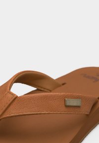 Καφέ δερμάτινο σανδάλι τύπου φλιπ-φλοπ με υφασμένη λουρίδα και μεταλλική πλακέτα με λογότυπο στη μία πλευρά, πάνω σε λευκό φόντο.