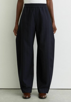 Person iført sorte bredbenede bukser med nålestriber og elastisk talje samt brune sko, stående foran en lys, ensfarvet væg.