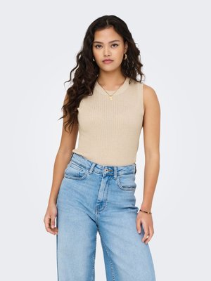 Jeune femme aux longs cheveux bouclés portant un haut sans manches beige et un jean taille haute bleu clair sur un fond uni.