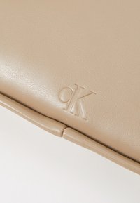 Beżowa skórzana saszetka z wytłoczonym logo "ck". Gładka faktura z delikatnym połyskiem, z designem typu fold-over i zaokrąglonymi krawędziami.