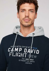 Sudadera navy con capucha gris, que presenta texto impreso en blanco y azul. El texto incluye coordenadas y el diseño "CAMP DAVID FLIGHT CREW".