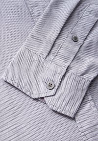 Camicia grigia chiara con un motivo testurizzato. Presenta un colletto a punta, dettagli con bottoni e cuciture visibili lungo il polsino.