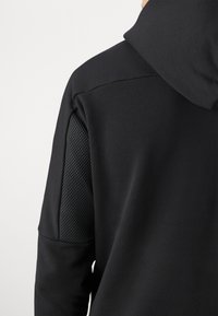 Felpa nera con un pannello a rete testurizzato sulla spalla sinistra. Tessuto liscio con una vestibilità comoda e cappuccio regolabile.