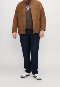 Veste en daim marron avec un col montant sur un t-shirt sombre arborant un motif floral, associée à un jean foncé et des baskets blanches avec des accents noirs.