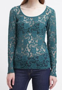 Teal spetslongärmad topp med blommönster och genomskinlig kropp samt rundad halsringning, matchad med mörka jeans.