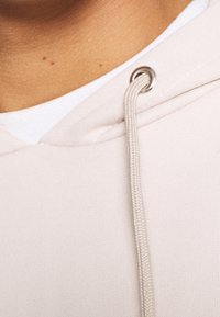 Felpa beige con cappuccio e lacci, dotata di occhielli argentati. Il materiale ha una texture liscia, con una camicia bianca a strati visibile sotto.