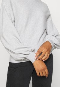 Pull en maille grise avec manches bouffantes et poignets côtelés, associé à un denim foncé. Le tissu semble doux avec une texture subtile.