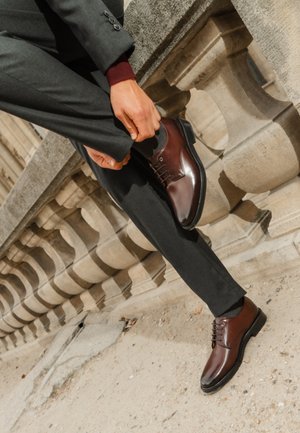 Scarpa da vestir in pelle marrone con punta arrotondata, design con lacci e finitura lucida. Indossata con pantaloni grigi scuri su una balaustra di pietra.