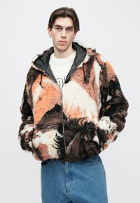 Giacca con zip in pile con un mix di motivi arancioni, marroni e crema, dotata di cappuccio, tasche laterali e finitura morbida e testurizzata.