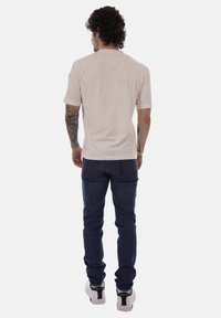 Uomo con tatuaggi, capelli ricci, che indossa una maglietta beige, jeans blu scuro e sneakers bianche, in piedi di spalle su sfondo bianco.