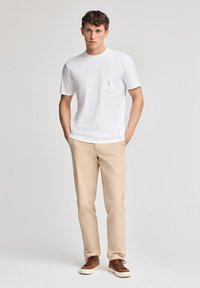 T-shirt bianco a maniche corte con taschino, abbinato a pantaloni beige chiaro e sneakers marroni. Tessuto morbido, vestibilità rilassata, design minimal.