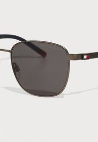 Occhiali da sole con lenti scure e quadrate, montatura in metallo sottile color bronzo e aste navy. Presentano un piccolo accento rosso e blu sul lato.