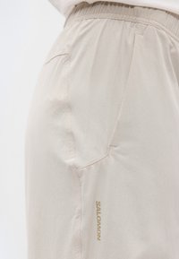 Μπεζ σορτς με ελαστική μέση, πλαϊνή τσέπη και το λογότυπο "Salomon" τυπωμένο κατακόρυφα κοντά στη ραφή της τσέπης.