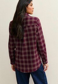 Camisa de cuadros burdeos de botones con mangas largas, que presenta líneas crema y un dobladillo curvado. El material parece suave con un acabado texturizado.