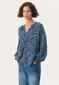 Blouse à motif floral dans des nuances de bleu et blanc, avec un col en V, des manches longues bouffantes et un tissu doux et texturé. Coupe et design décontractés.