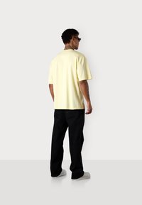 Gelbes kurzärmliges T-Shirt aus Baumwolle; getragen mit schwarzen lockeren Hosen und weißen Turnschuhen aus der Rückansicht.