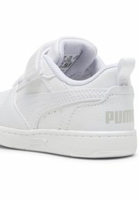 Bílé tenisky Puma s hladkým syntetickým svrškem, suchým zipem, polstrovaným límcem a texturovanou podrážkou. Na patě je decentní logo.