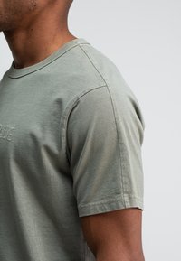 Close-up van een persoon die een lichtgroen T-shirt met korte mouwen draagt, met gestikte details op de borst, tegen een effen lichte achtergrond.