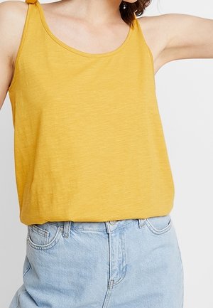 Débardeur sans manches jaune moutarde avec bretelles nouées, porté avec un jean denim bleu clair taille haute, raccourci au milieu du torse.