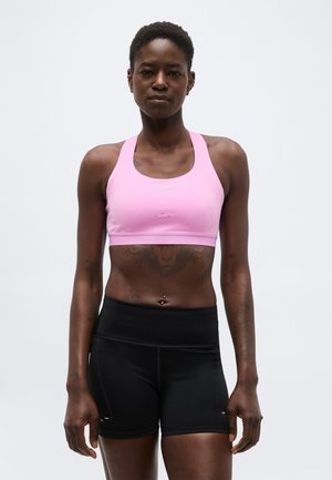 Femeie atletică în picioare, purtând un sutien sportiv roz și pantaloni scurți sportivi negri cu talie înaltă, pe un fundal simplu, deschis la culoare.