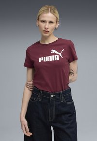 Bordo pamučna majica s bijelim "PUMA" logotipom i grafikom skakačkog pumasa. Okrugli dekolte, kratki rukavi, uparena s tamnim trapericama visokog struka.