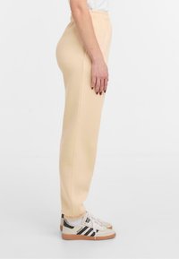 Beige Sweatpants mit elastischem Bund, schmal zulaufenden Säumen, seitlichen Taschen und einer glatten Textur, kombiniert mit weißen und schwarzen Sportschuhen.