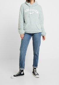 Ljusblå fleecehuva med en kenguruficka och vit "HOLLISTER"-logotyp. Parat med uppvikta denimjeans och svarta högskaftade sneakers.