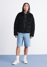 Giacca a vento nera con zip frontale, caratterizzata da sezioni trapuntate. Indossata con pantaloni corti in denim azzurro e sneakers crema. Sfondo bianco.