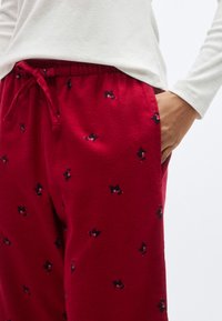Pantalon de pyjama rouge avec une taille à cordon, présentant une texture douce et de petits motifs de chiens noirs partout. Poche latérale visible.