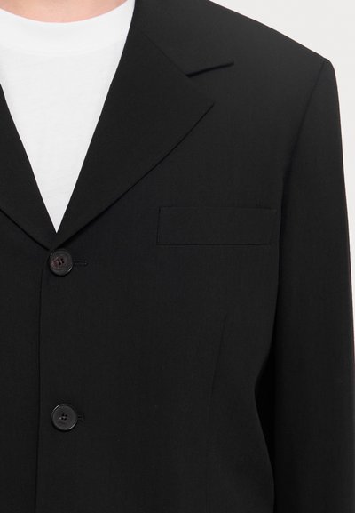 Schwarzer Blazer aus glattem Stoff, mit einem Reverskragen, zwei Knöpfen und einer aufgesetzten Tasche auf der linken Seite.