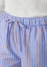 Pantalons rayés bleu clair avec des lignes verticales rouges et blanches. Taille élastique avec cordon de serrage. Tissu doux et texturé.