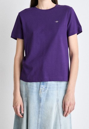 Femme portant un t-shirt violet uni à manches courtes avec un petit logo sur la poitrine et une jupe en jean bleu clair avec des coutures à l'avant.