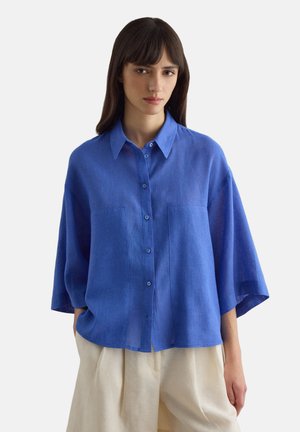 Jonge vrouw die een loszittend blauw overhemd met wijde mouwen en beige geplooide broek draagt, staand met handen in de zakken.