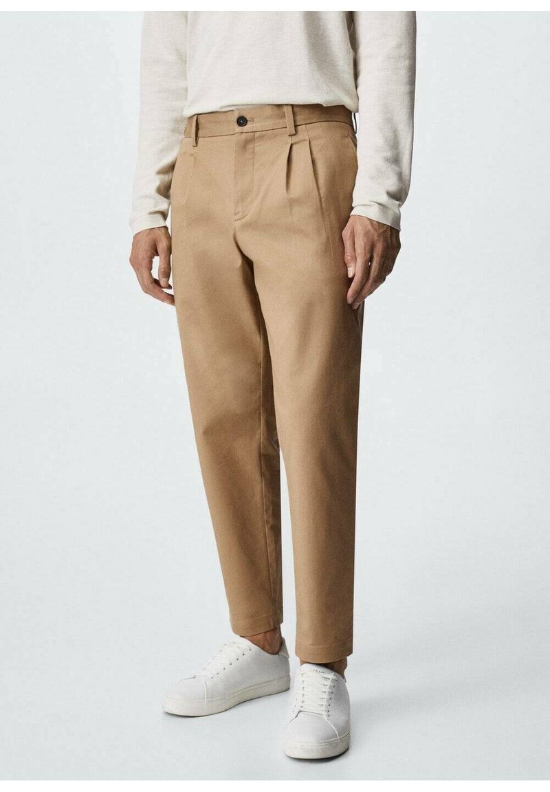 Homme portant un pantalon plissé beige, une chemise blanche à manches longues et des baskets blanches basses, debout sur un fond uni clair.
