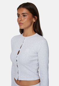 Light gray ribbed long-sleeve top with a round neckline, front button closure, and subtle embroidered logo detail. Cropped length.