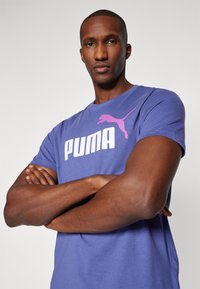 Violeta kokvilnas T-krekls ar izteiksmīgu baltu "PUMA" logo un rozā pumju grafiku. Īsas piedurknes, apaļa kakla izgriezums un brīva piegriezuma dizains.