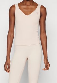 Camiseta de tirantes de color beige claro con canalé, escote en V, tirantes anchos y un pequeño logo triangular de marca en el lado izquierdo. Textura suave.