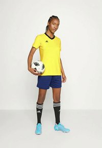 Gelbes Fußballtrikot mit schmalem schwarzen Kragen, blaue Shorts, schwarze Socken mit weißen Streifen und blaue Fußballschuhe. Hält einen schwarz-weißen Fußball.