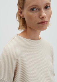 Primo piano di una donna dalla pelle chiara e capelli biondi corti, che indossa un maglione a costine color crema, guardando dolcemente verso la fotocamera.
