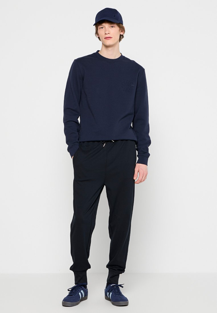 Marine sweatshirt met een ronde hals en klein logo; gecombineerd met zwarte joggingbroeken met elastische boorden. Een marine pet en sneakers maken de look compleet.
