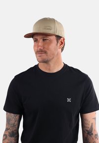 La casquette présente un corps marron clair, une visière plate marron foncé et un logo brodé. Le modèle porte un t-shirt noir avec un petit motif blanc.